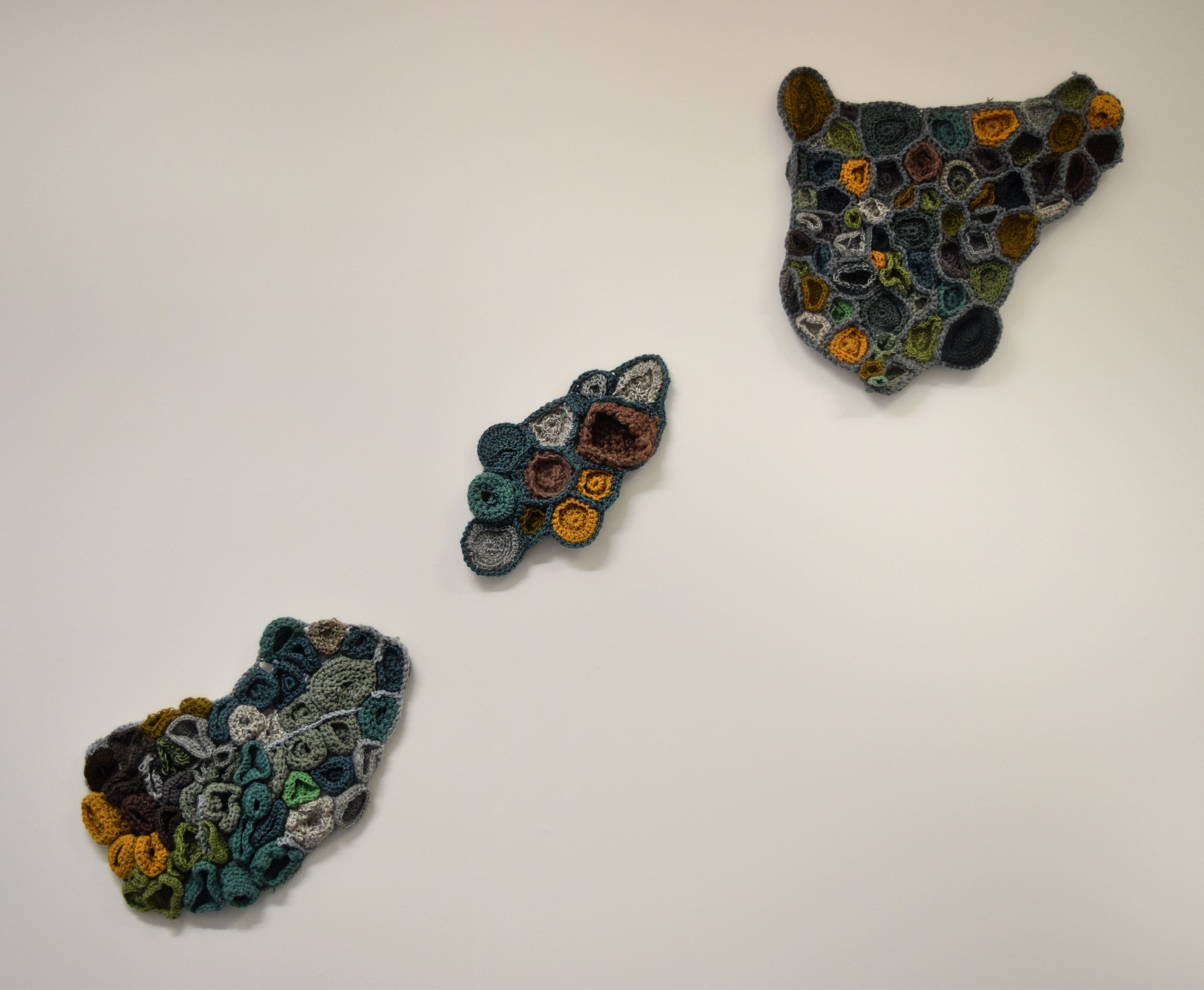 3 highly textured crochet sculptures hanged diagonally on a white wall, all 3 in natural colours of green, yellow and grey