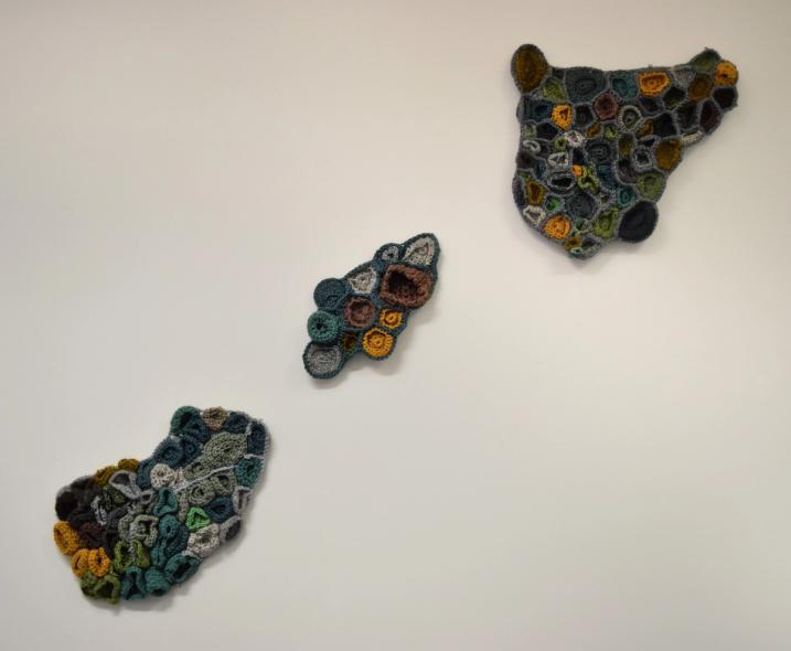 3 highly textured crochet sculptures hanged diagonally on a white wall, all 3 in natural colours of green, yellow and grey