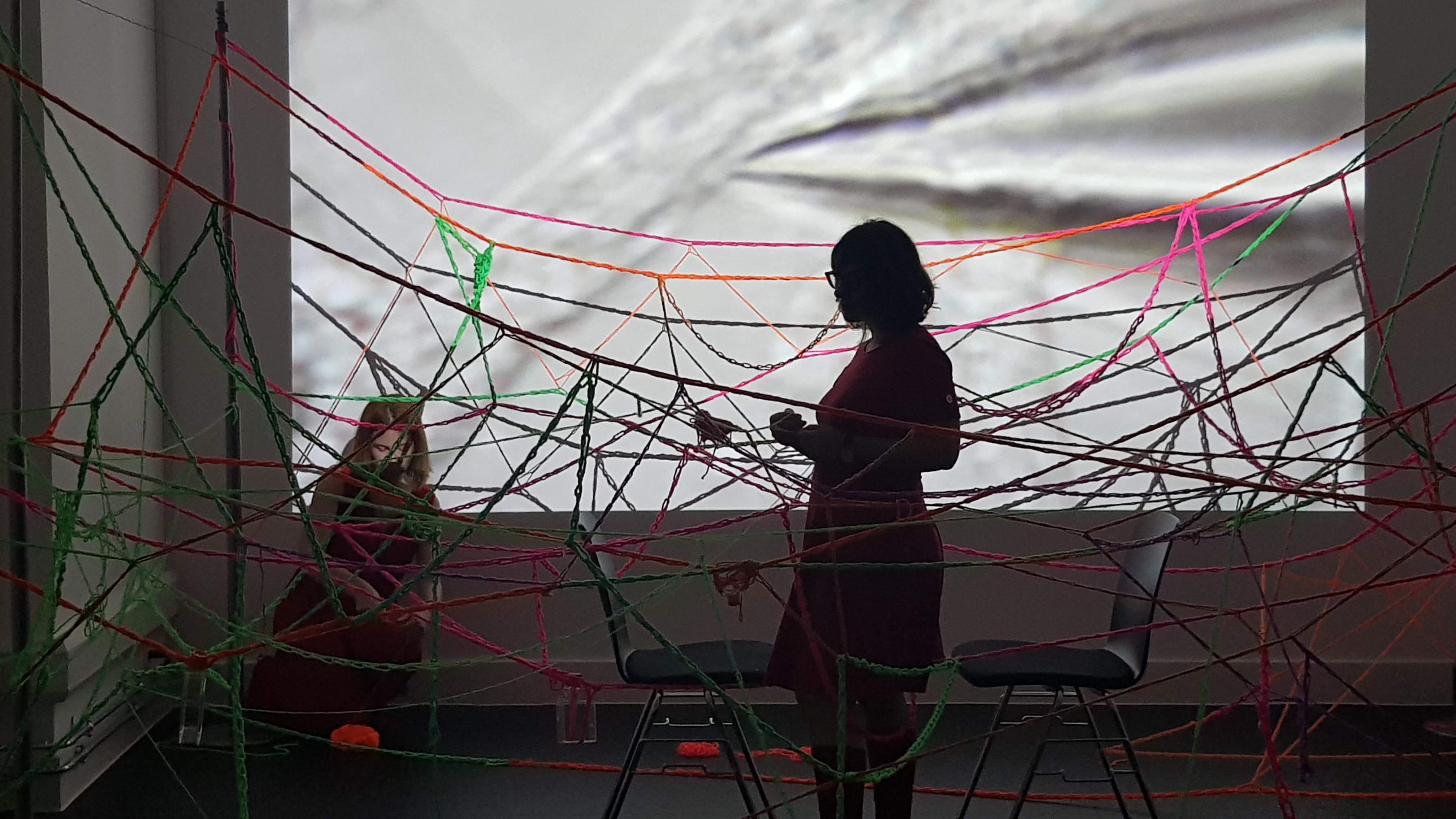 Artists Barbara Touati-Evans and Susan Merrick making a wool web in front of neuroscience video