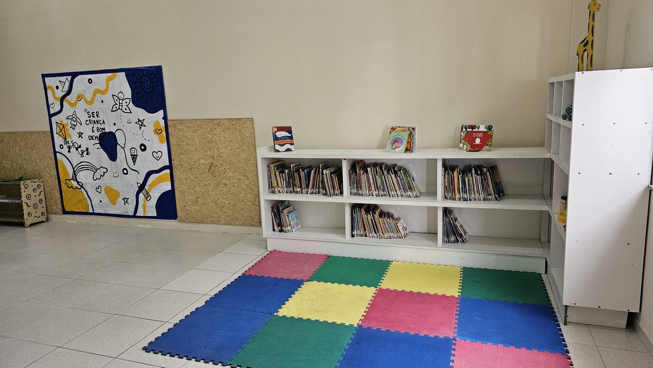 Ateliê e biblioteca infantil com livros, piso emborrachado e painel com a frase “Ser criança é bom demais”