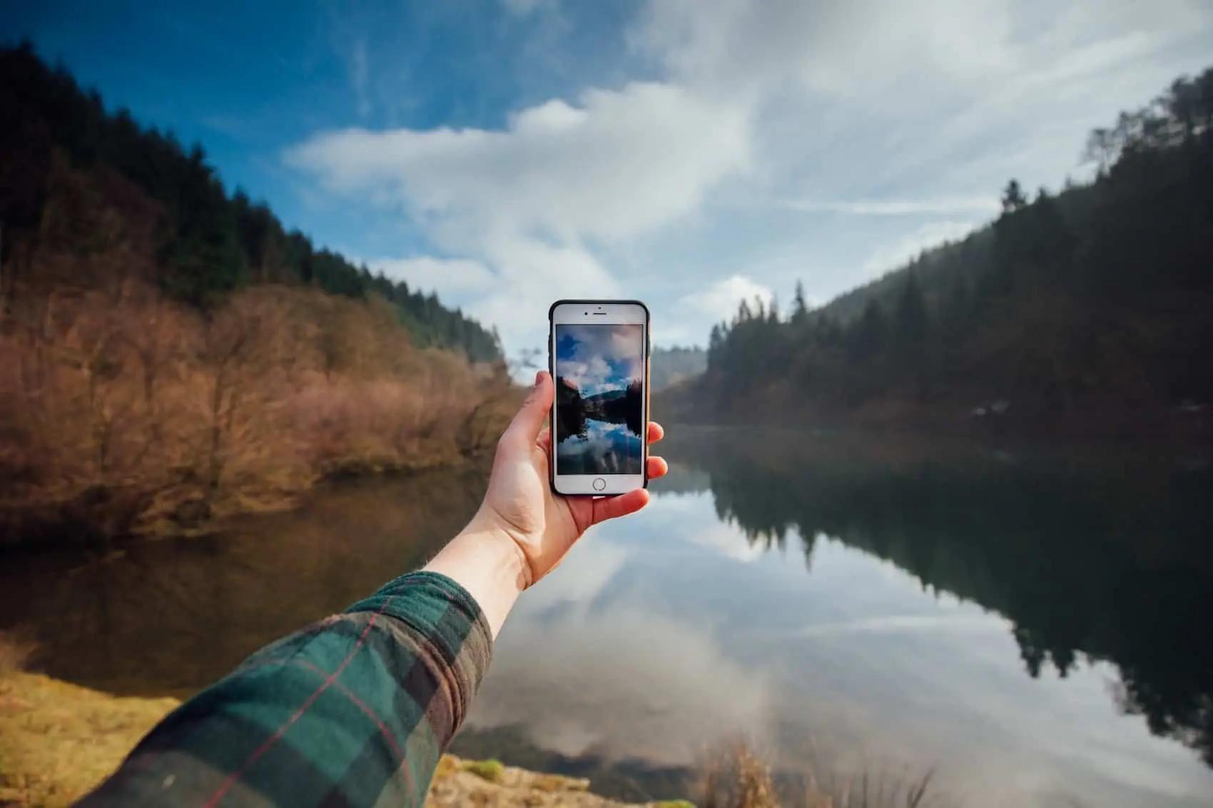 A hand holds up a smartphone to photograph a still lake surrounded by forested hills