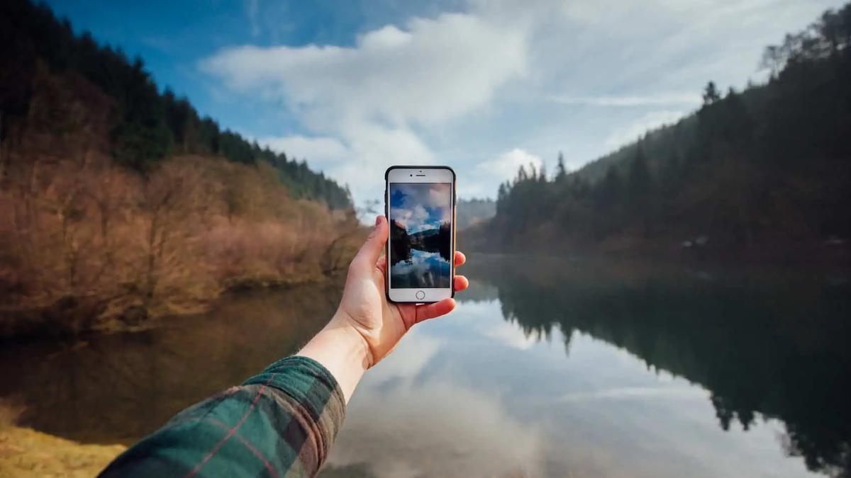 A hand holds up a smartphone to photograph a still lake surrounded by forested hills