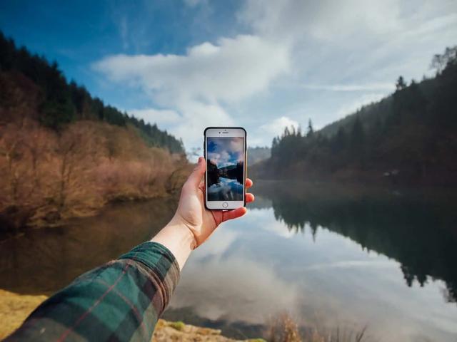 Una mano sostiene un teléfono para fotografiar un lago tranquilo rodeado de colinas boscosas