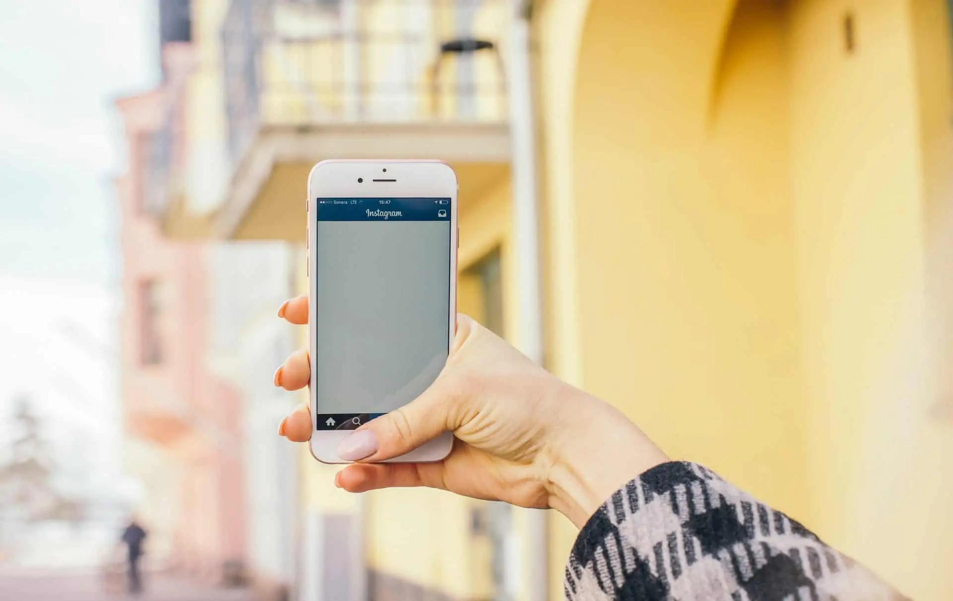 A hand holds up a phone showing the Instagram app open, with a yellow building in the background