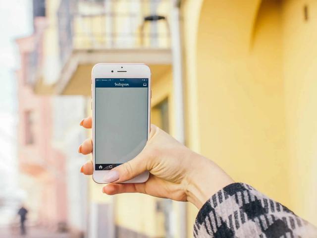 A hand holds up a phone showing the Instagram app open, with a yellow building in the background