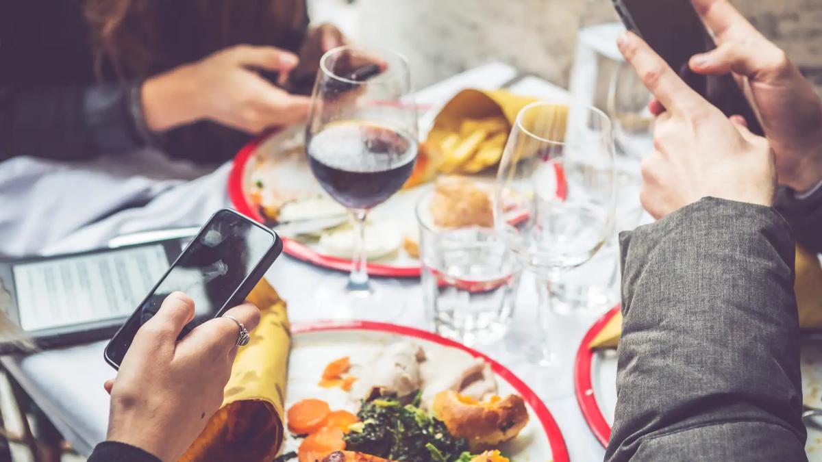 Three people at a restaurant table, each holding a smartphone. Plates of food, wine, and water glasses are visible.