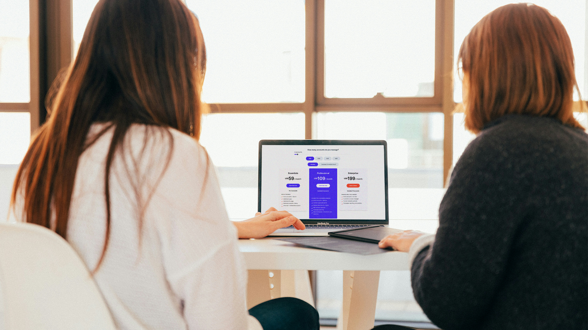 Two people viewing a laptop screen showing a pricing page with Essentials $59, Professional $109, and Enterprise $199 plans