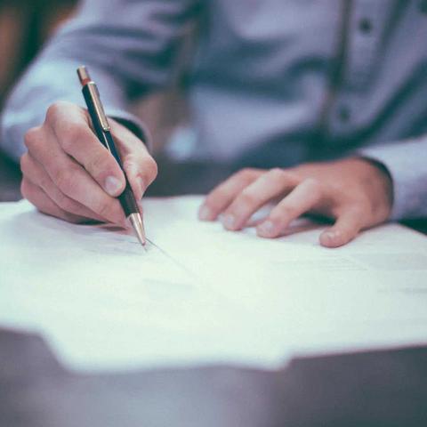 Person in a blue shirt writing on white paper with a black and gold pen. Dark, serious mood.