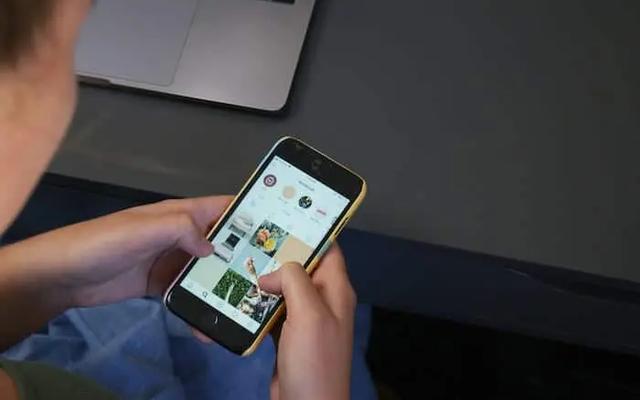 Person holding a phone scrolling an Instagram grid, with a laptop in the background