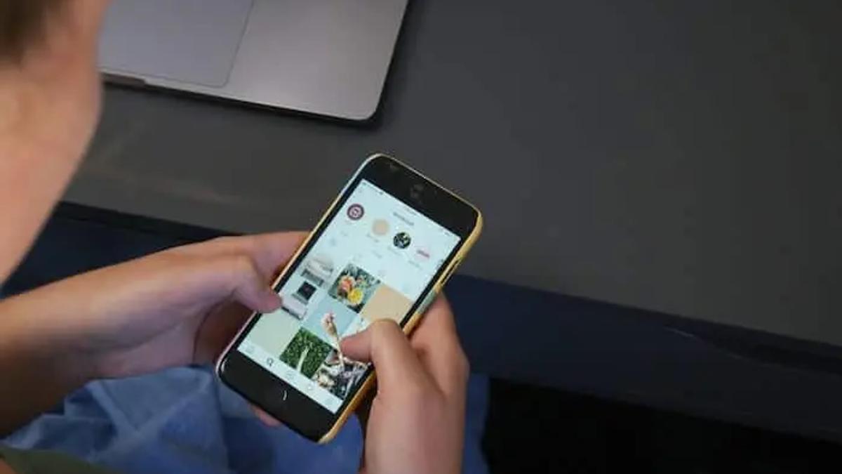 Person holding a phone scrolling an Instagram grid, with a laptop in the background