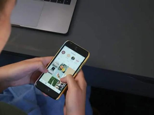 Una persona sostiene un teléfono mostrando una cuadrícula de Instagram, con una laptop al fondo