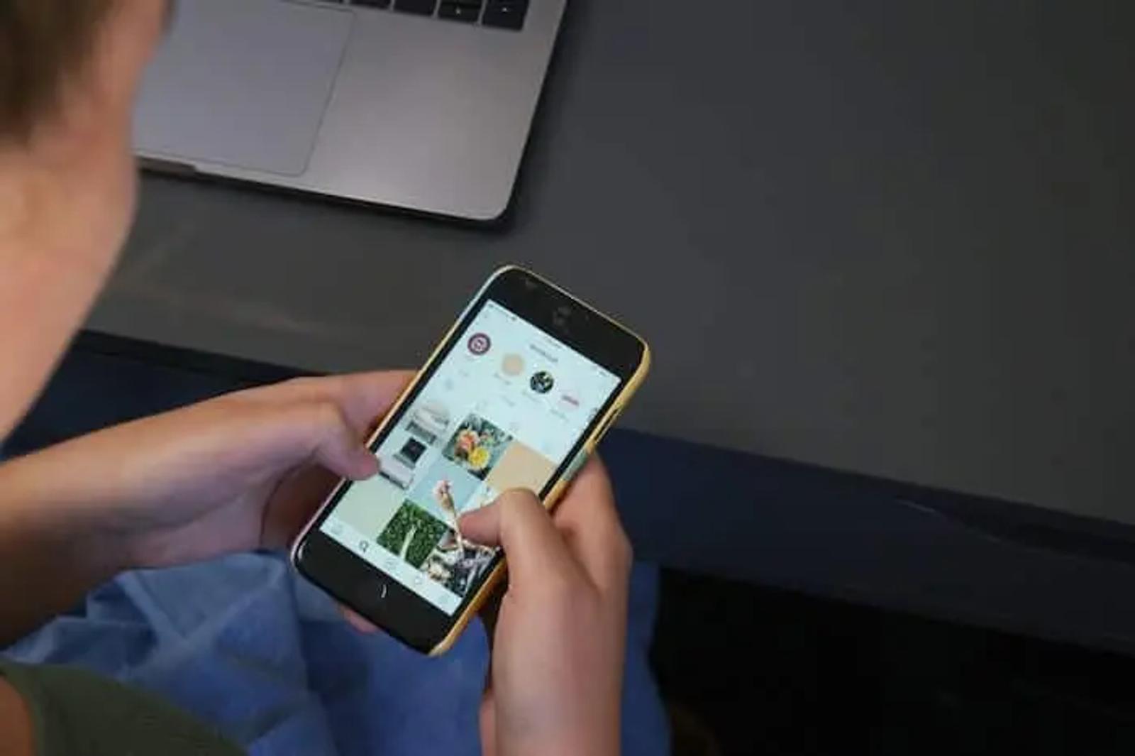 Una persona sostiene un teléfono mostrando una cuadrícula de Instagram, con una laptop al fondo