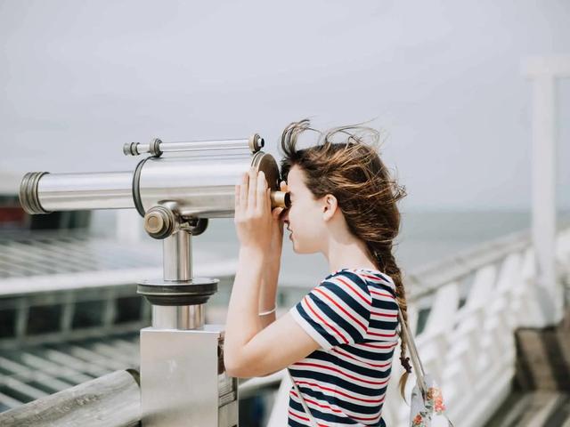 Young woman with brown hair in a striped shirt looking through a silver telescope by the ocean on a sunny day