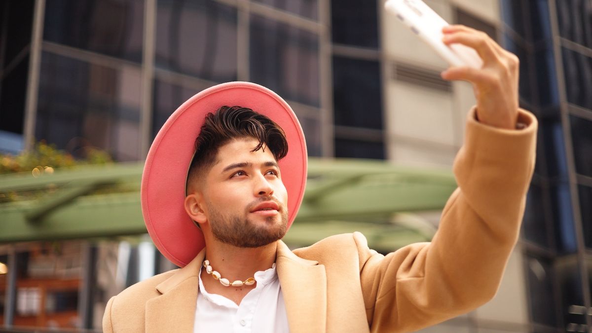 Man in a pink hat, tan coat, and shell necklace taking a selfie with a white phone in an urban setting
