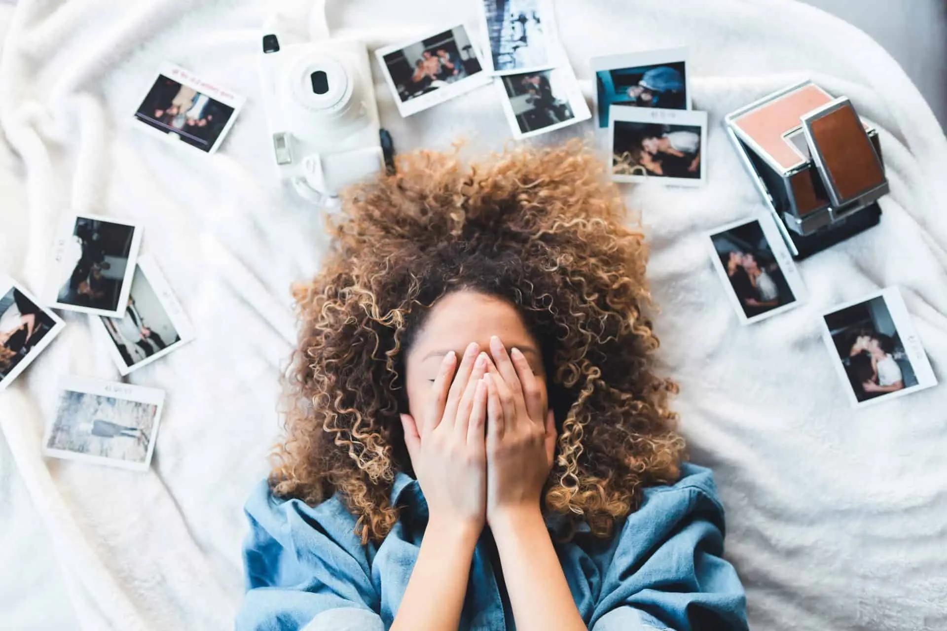 Woman with curly hair lying on a bed, covering her face, surrounded by scattered polaroid photos and a camera
