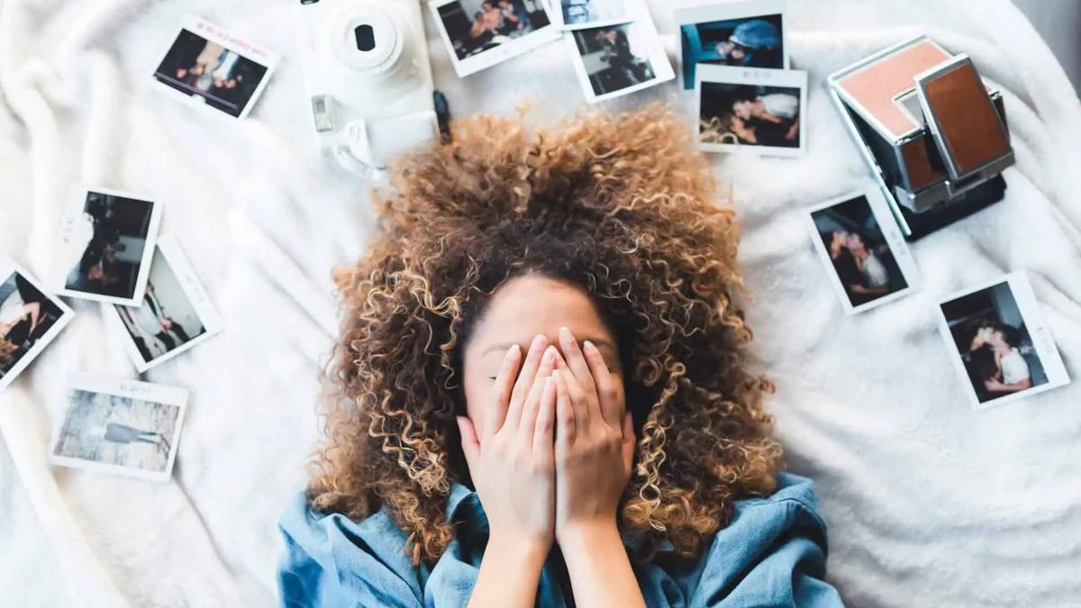 Woman with curly hair lying on a bed, covering her face, surrounded by scattered polaroid photos and a camera