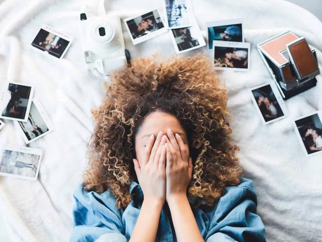 Mujer de cabello rizado acostada cubriéndose el rostro, rodeada de fotos polaroid y una cámara instantánea