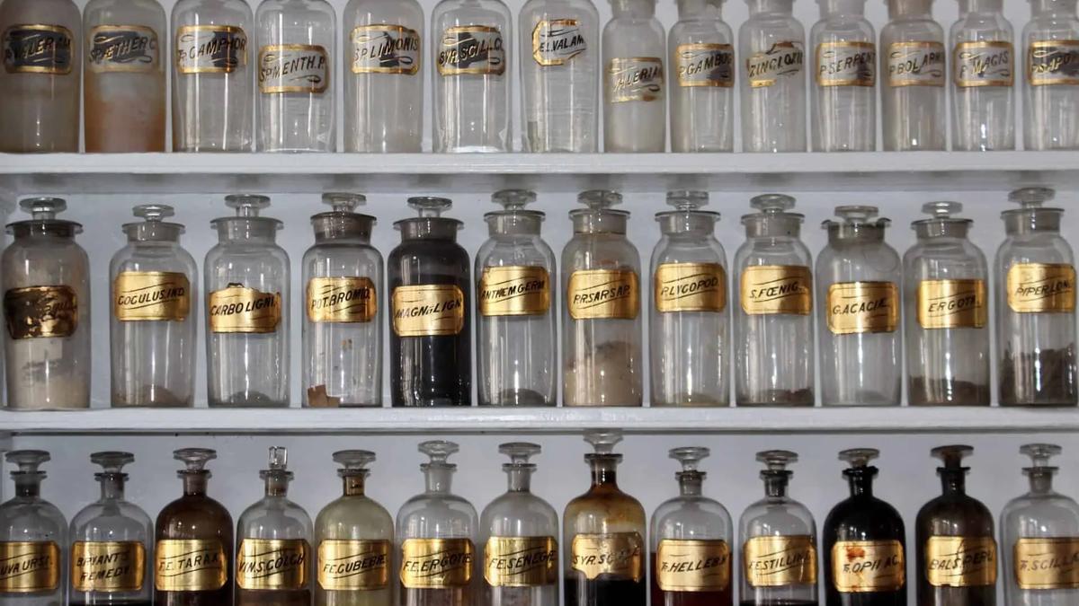 Three shelves of antique apothecary bottles with glass stoppers and gold labels, some with contents, some empty.