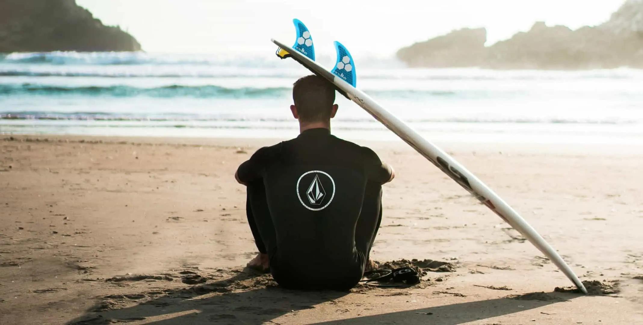 Una persona con traje de neopreno sentada en la arena frente al mar junto a una tabla de surf