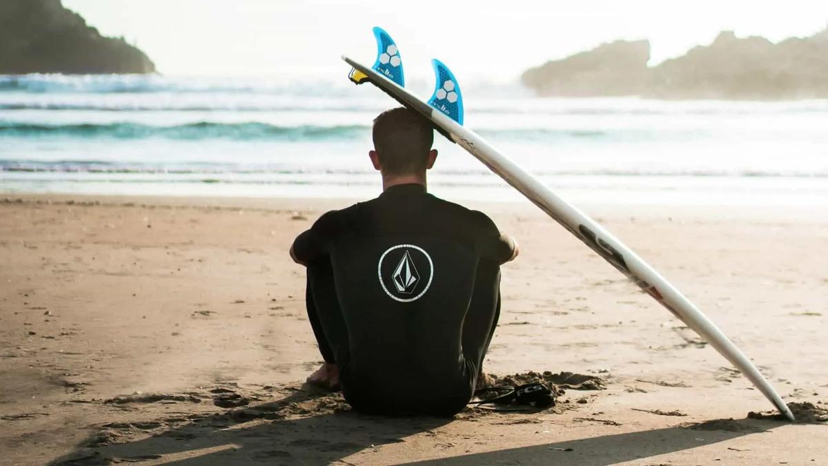 A person in a wetsuit sits on the sand facing the ocean beside an upright surfboard