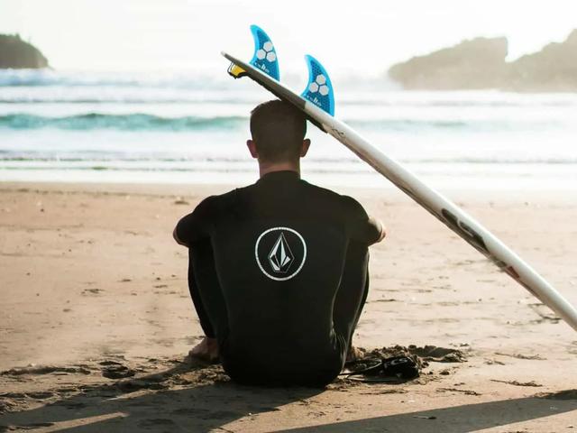 Una persona con traje de neopreno sentada en la arena frente al mar junto a una tabla de surf