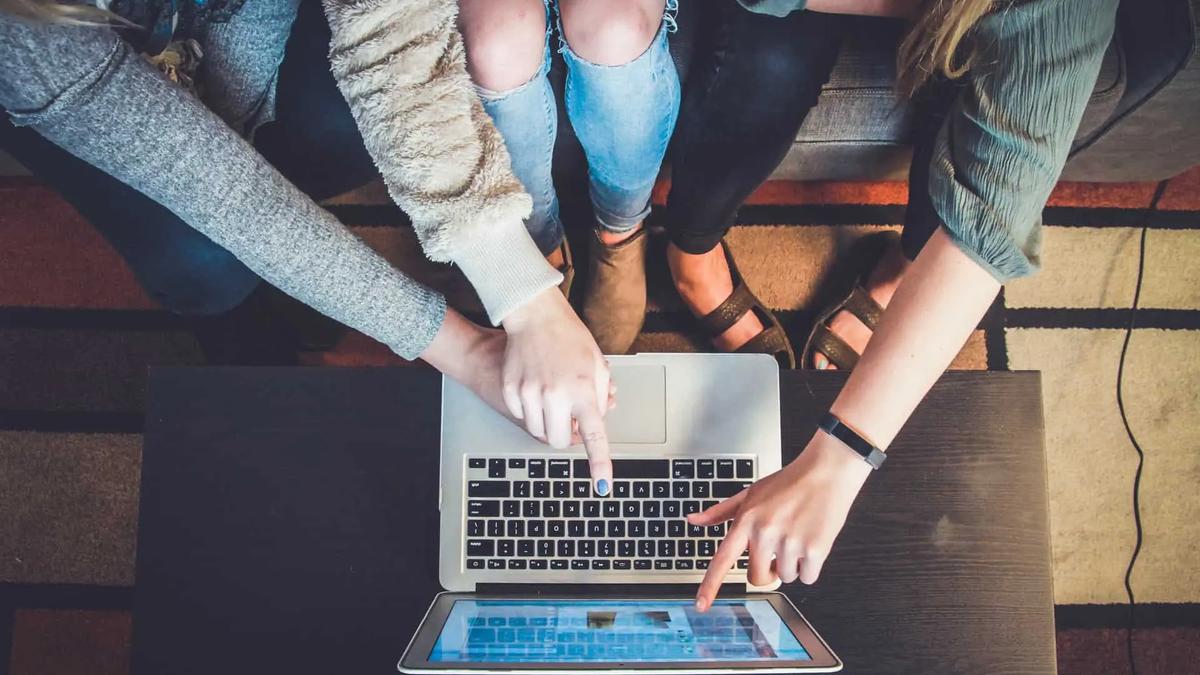 Tres personas señalando una laptop y una tablet sobre una mesa oscura, con una alfombra colorida al fondo. Ambiente colaborativo.