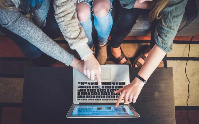 Three people pointing at a laptop and tablet on a dark table, with a colorful rug in the background. Collaborative mood.