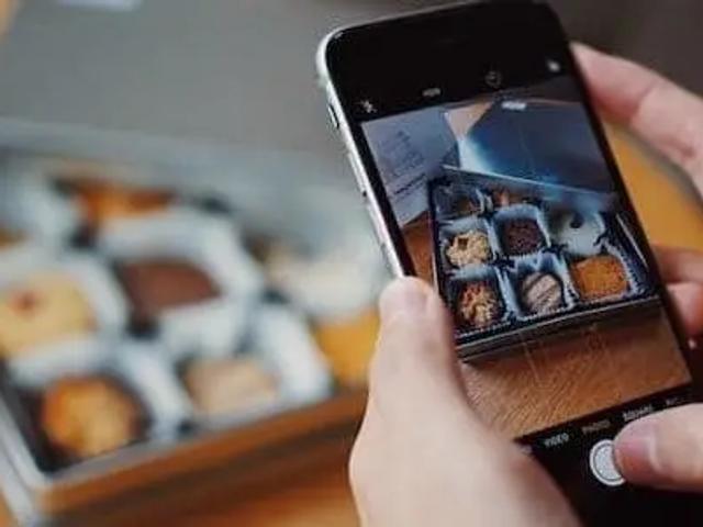 Manos sosteniendo un teléfono para fotografiar una caja de galletas surtidas sobre una mesa de madera