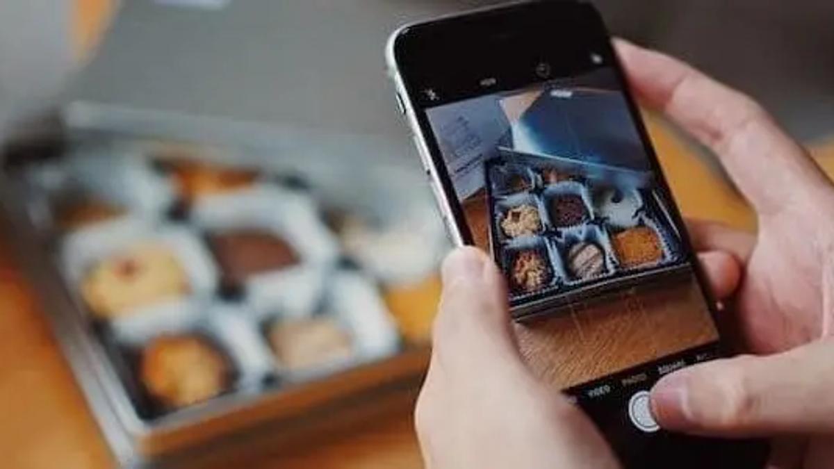 Hands holding a smartphone photographing a box of assorted cookies on a wooden table