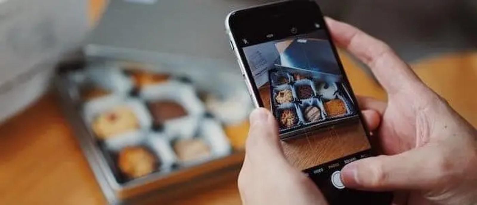 Manos sosteniendo un teléfono para fotografiar una caja de galletas surtidas sobre una mesa de madera