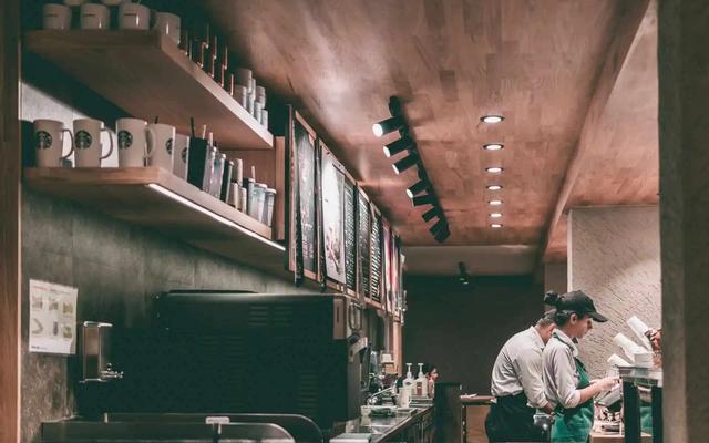 Empleados trabajando detrás del mostrador de una cafetería, con tazas y cartas de menú en los estantes