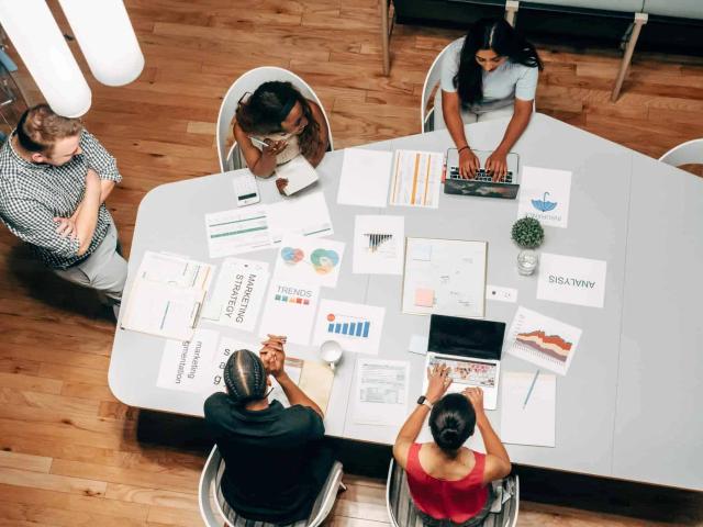 Overhead view of a team meeting around a table covered with charts, laptops and marketing strategy papers