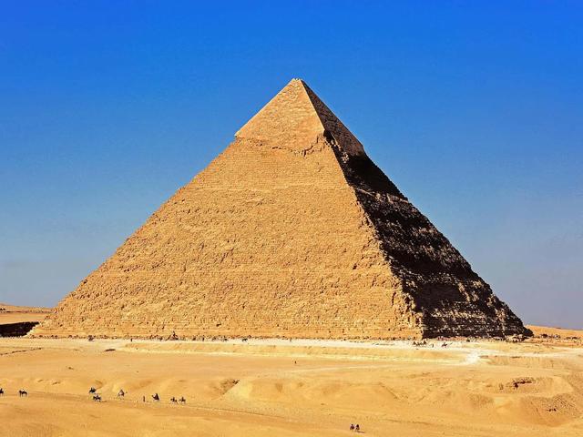 Great Pyramid of Giza in a desert landscape under a clear blue sky. Small figures and camels are visible.