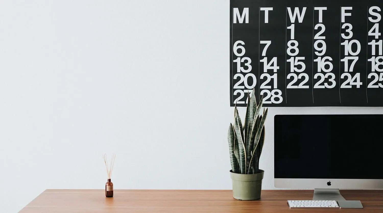 Un calendario de pared sobre un escritorio con una planta, un difusor de aromas y un ordenador