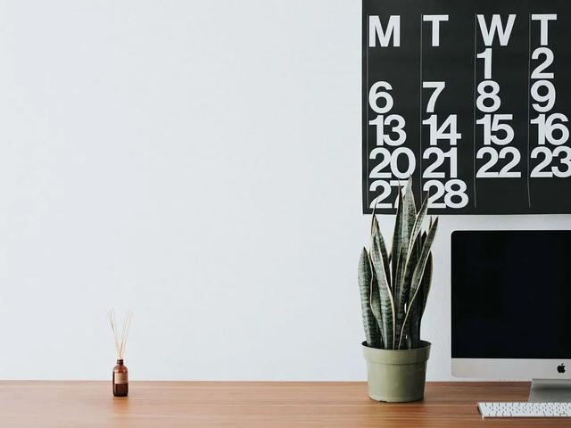 Un calendario de pared sobre un escritorio con una planta, un difusor de aromas y un ordenador
