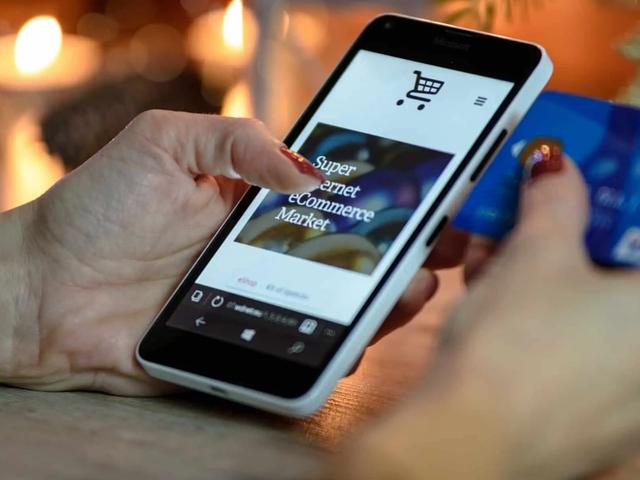 A hand holding a phone showing a shopping cart icon on an ecommerce site, while the other hand holds a credit card