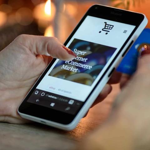 Una mano sostiene un telefono con un icono de carrito de compras, mientras la otra sostiene una tarjeta de credito