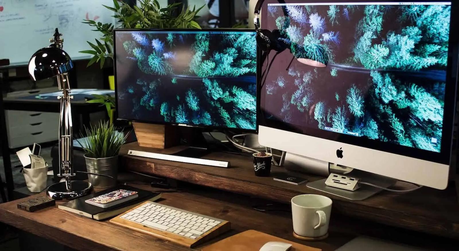 Un escritorio con dos monitores que muestran una imagen aérea de un bosque, teclado, lámpara y una taza de café