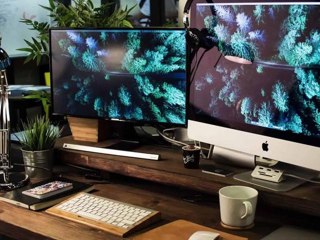 Un escritorio con dos monitores que muestran una imagen aérea de un bosque, teclado, lámpara y una taza de café
