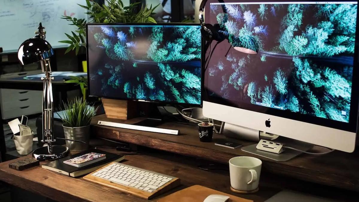 A desk with two monitors displaying an aerial forest image, a keyboard, lamp and coffee mug
