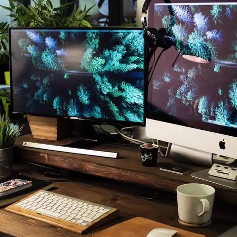 Un escritorio con dos monitores que muestran una imagen aérea de un bosque, teclado, lámpara y una taza de café