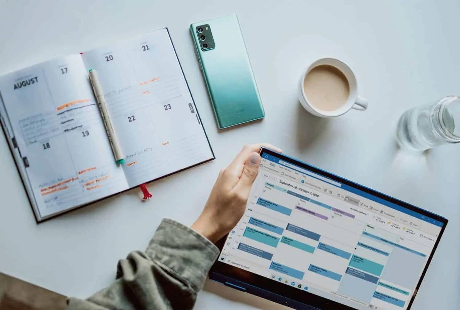 Una agenda de papel, un teléfono, un café y un portátil con un calendario semanal en la pantalla