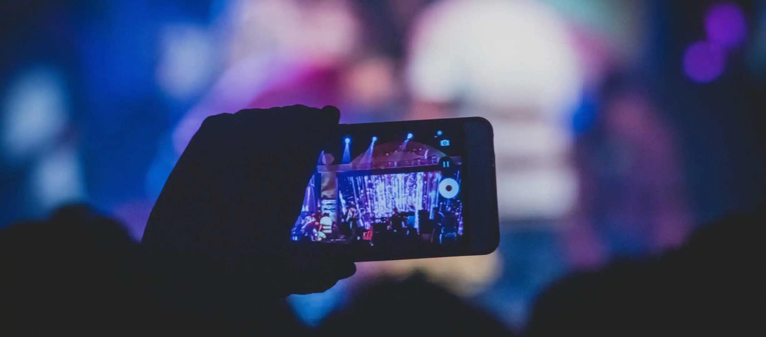 A hand holds up a phone recording video of a concert stage in a dark venue