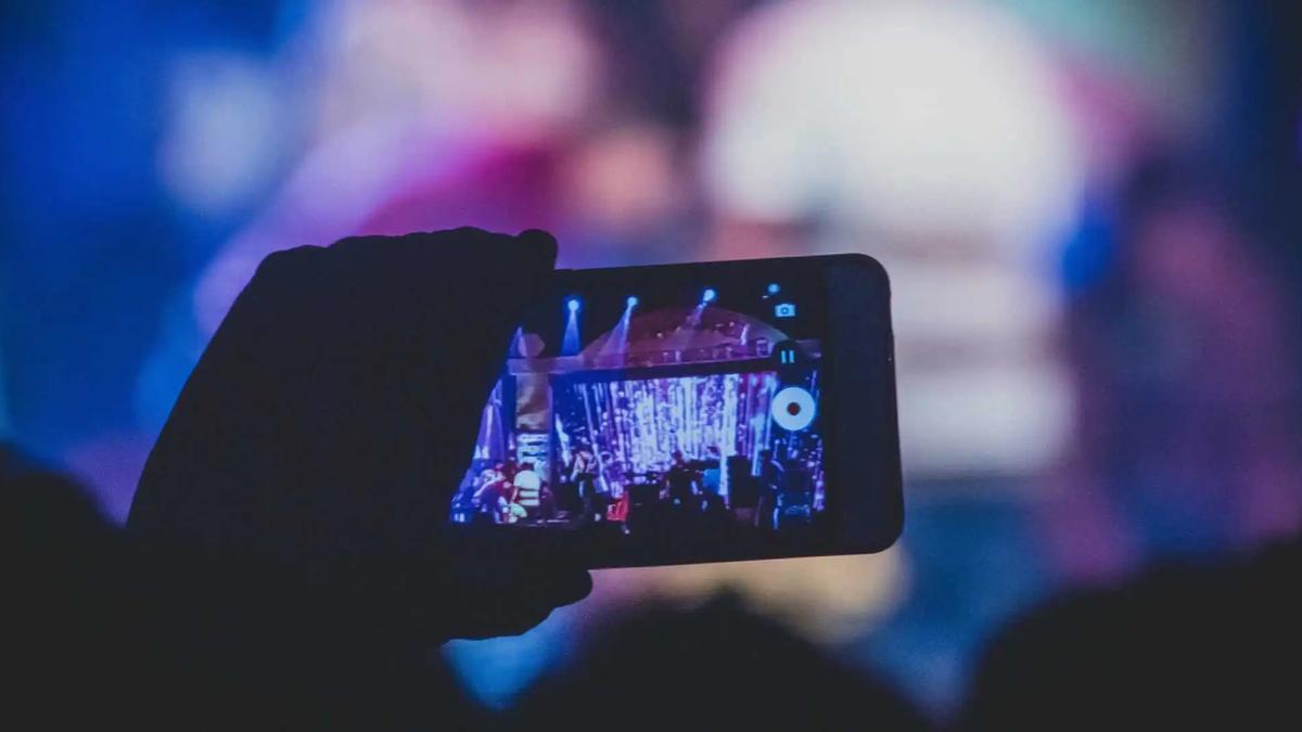 Una mano sostiene un teléfono grabando en video un escenario de concierto en un lugar oscuro
