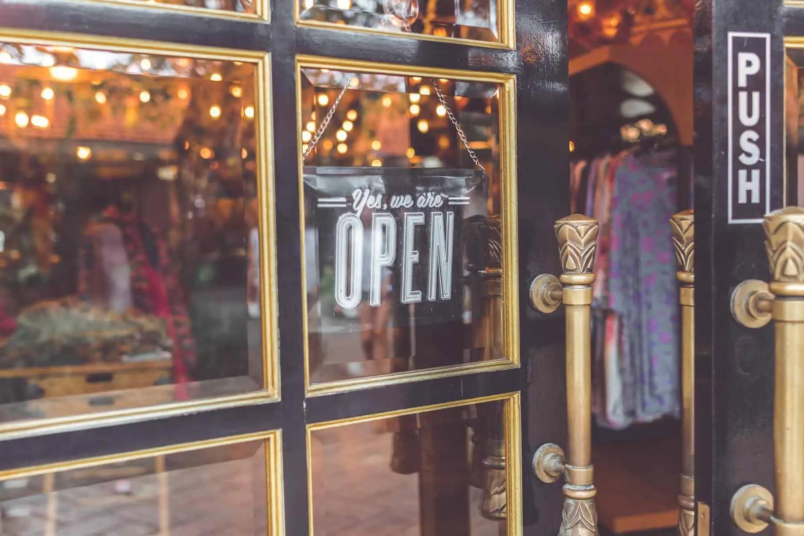 Puerta de vidrio de una tienda con cartel de abierto, manijas doradas y ropa visible en el interior