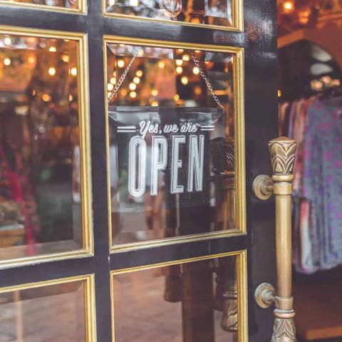 Puerta de vidrio de una tienda con cartel de abierto, manijas doradas y ropa visible en el interior