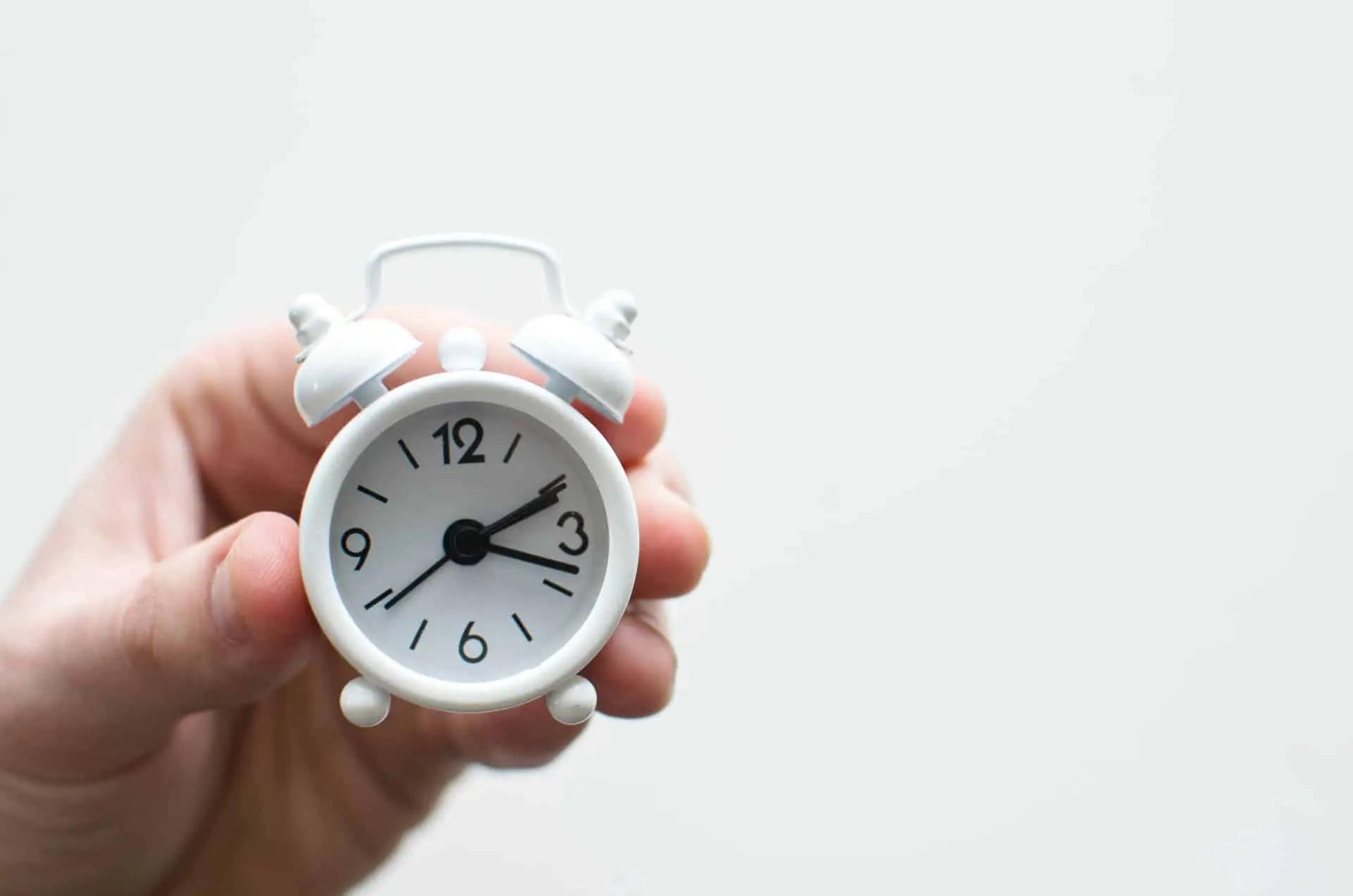 A hand holding a small white miniature alarm clock against a plain background