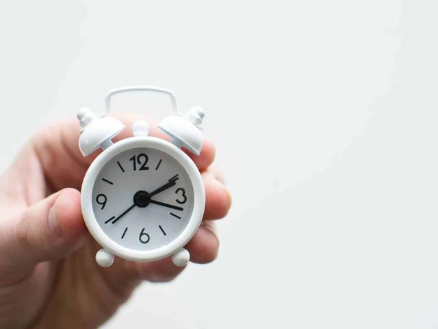 A hand holding a small white miniature alarm clock against a plain background