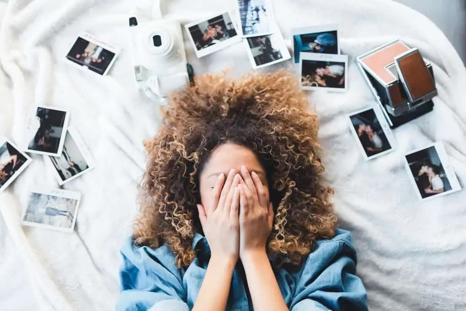 Mujer recostada cubriéndose el rostro, rodeada de fotos polaroid dispersas y una cámara instantánea