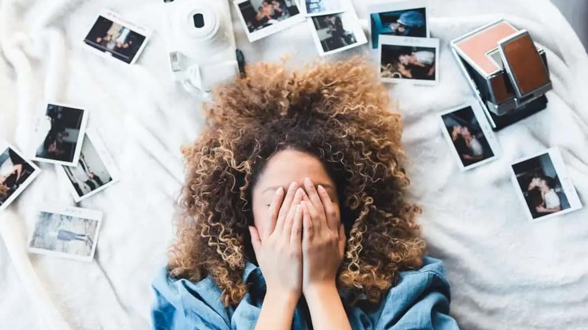 Mujer recostada cubriéndose el rostro, rodeada de fotos polaroid dispersas y una cámara instantánea